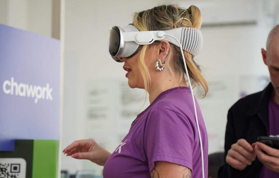 Mulher loira de camiseta roxa utiliza óculos de realidade virtual de alta tecnologia, em frente a um painel com o logotipo da Chawork.